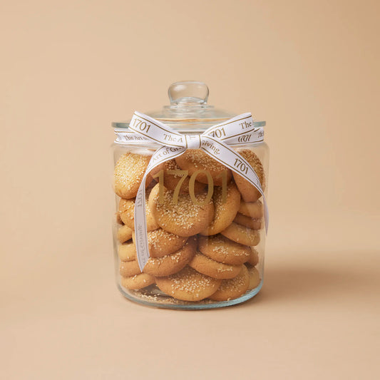 A glass jar filled with sesame seed shortbread rounds. The jar is tied with a ribbon and has a label with the name of the biscuits and the SANHA Halaal approved logo.