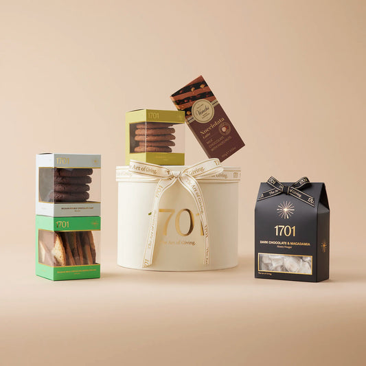The Cocoa Lovers Medium Gift Box is presented on a warm background. The gift box is a paper hat box. Inside the box, there are various chocolate treats, including handcrafted honey nougat, three boxes of delicious biscuits, and a bar of imported Italian chocolate. This gift box is perfect for chocolate lovers, whether shared or enjoyed alone.