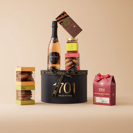 The Luxury Red Grape Juice Gift Box  is presented on a warm background. The gift box includes a box of handcrafted honey nougat, a selection of incredible biscuits, a jar of 1701 farm honey, a slab of imported Italian chocolate, and a bottle of Melozhori red grape juice. This gift box is perfect for those who appreciate celebrations without alcoholic drinks and the luxury of fine treats. The brown and gold design of the gift box adds a touch of elegance, making it perfect for any occasion or just because.