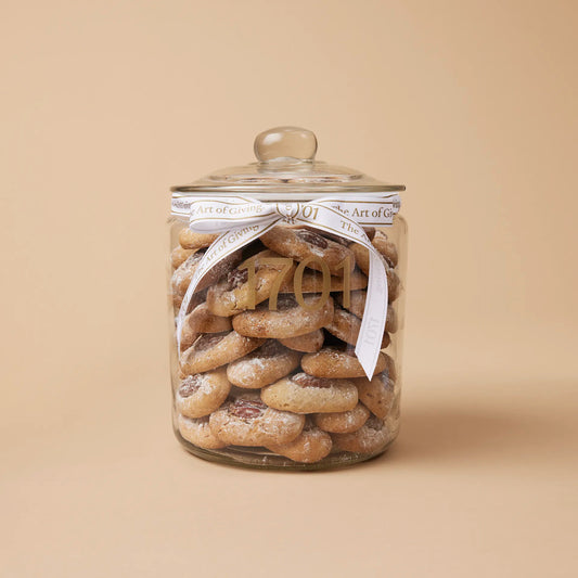 A 1kg glass jar of handmade biscuits with pecan nuts, presented in a glass jar with a hand-tied signature ribbon on a warm background, with the product label visible on the front of the jar. The biscuits are SANHA Halaal approved, making them a perfect gift idea for anyone who appreciates high-quality biscuits made with care.