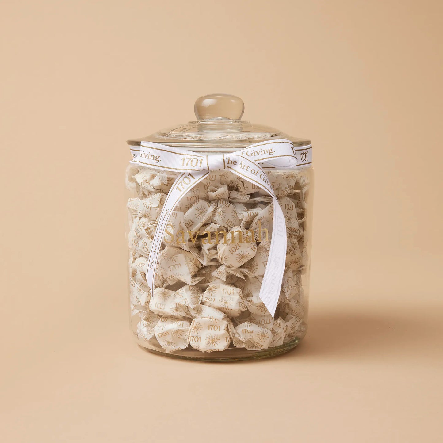 A glass jar filled with handmade honey nougat pieces, with a personalized label affixed to the front. The nougat is a blend of creamy white and golden brown colors, and the jar is finished with a signature handtied ribbon.