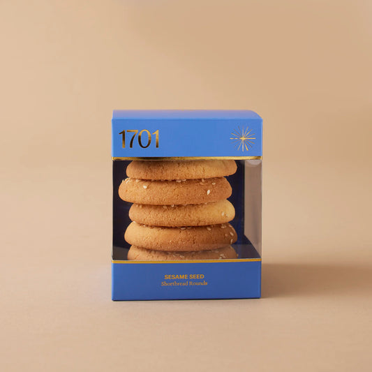 A box of Sesame Seed Shortbread Rounds (200g) with the product label visible on the front. The handmade biscuits are presented on warm background. The box is labeled as SANHA Halaal approved.
