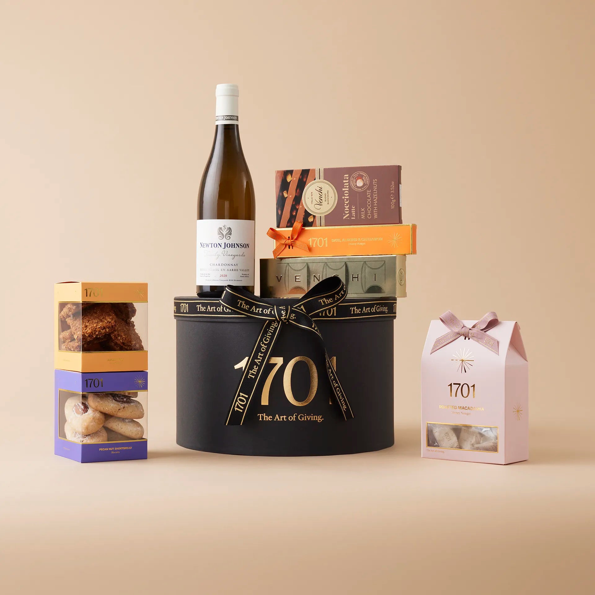 A gift box containing a bottle of Newton Johnson Family Vineyards 2020 Chardonnay, a box of handcrafted honey nougat, a nougat bar, two boxes of biscuits, and two imported Italian chocolates by Venchi. The box is decorated with a ribbon and placed on a wooden table with a warm background.
