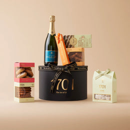 A gift box filled with South African treats, including a box of handcrafted honey nougat, a nougat bar, a selection of biscuits, imported Italian chocolate, and a bottle of Colmant MCC Non-Reserve or Rosé, presented in a hat box with a ribbon.