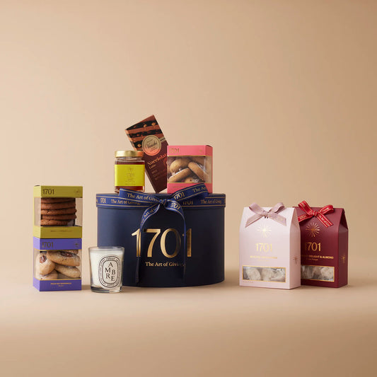 "Luxury Home" gift box featuring a large scented Diptyque candle and a selection of gourmet treats including handcrafted honey nougat, biscuits, farm honey, and imported Italian chocolate by Venchi. The gift box is presented in a signature hat box and is shown on a warm background. The gift box is tied with a ribbon, and a gift tag is visible on the top of the box.